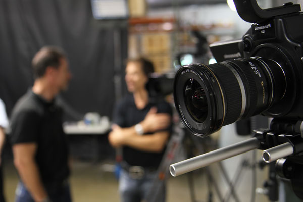 A photo of a video camera with two people in conversation behind the scenes